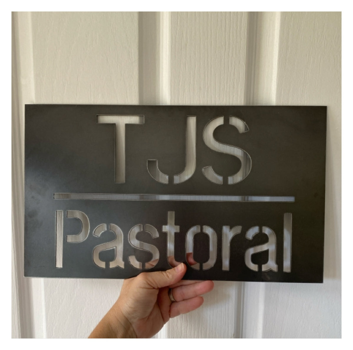 Metal sign with 'TJS Pastoral' text held against a neutral background
