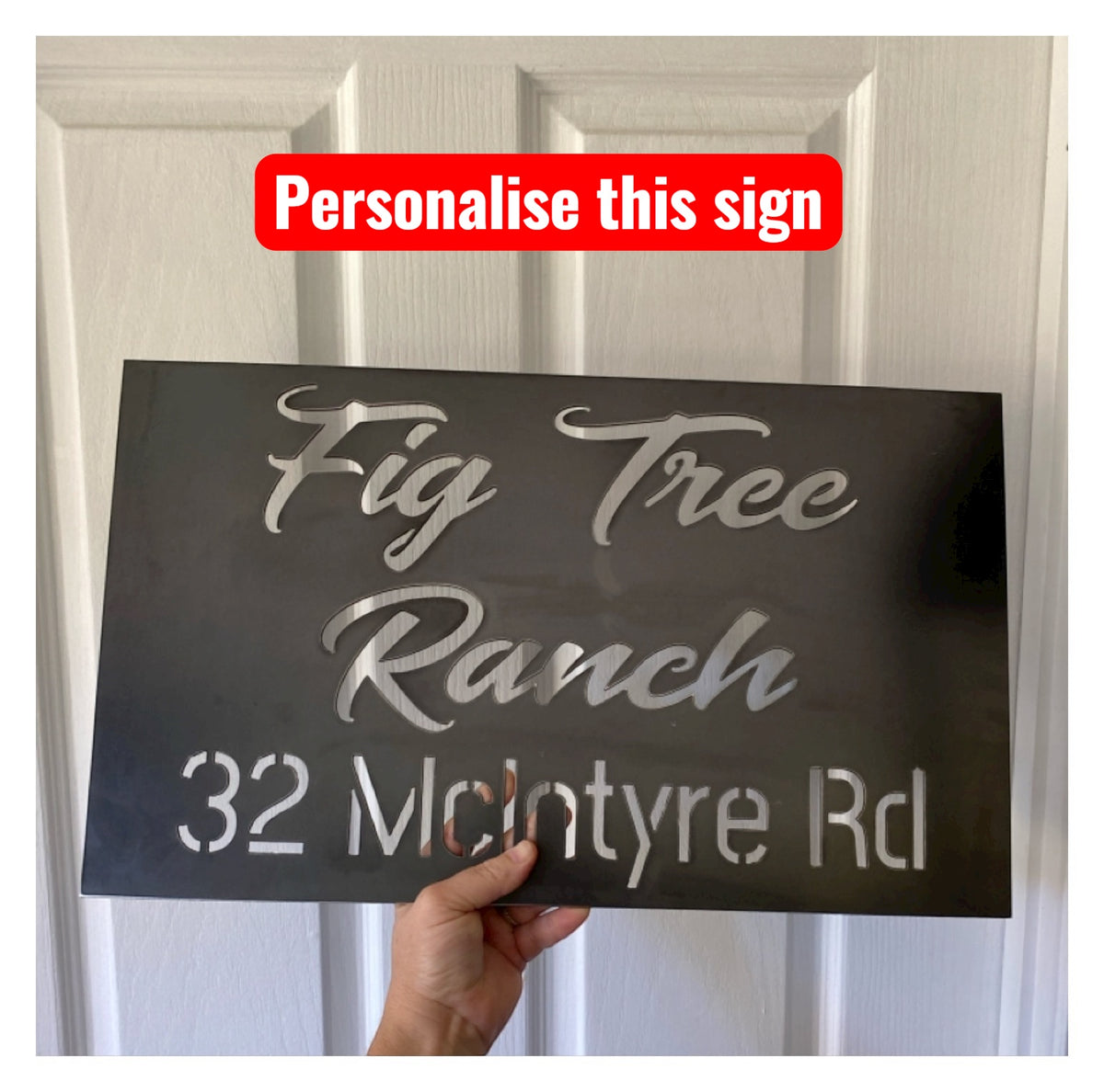 Hand holding a personalized address sign with text 'Fig Tree Ranch 32 McIntyre Rd' against a white background.