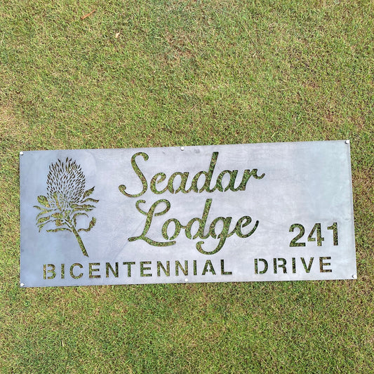 Metal sign for Seadar Lodge on Bicentennial Drive with a decorative plant design, placed on grass.