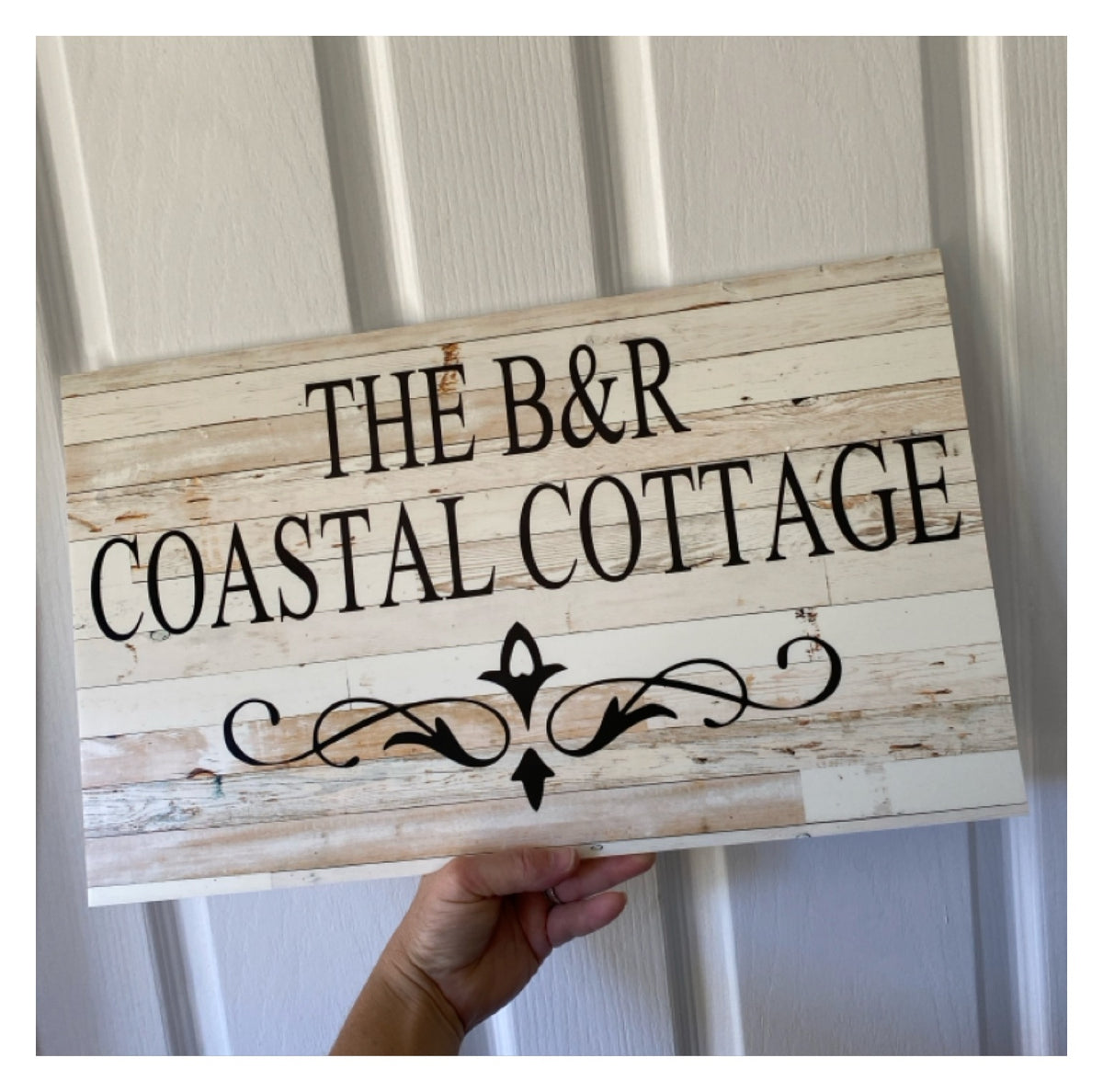 Wooden sign with 'The B&R Coastal Cottage' text held by a hand against a white background