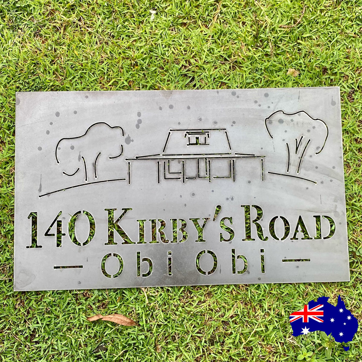 Metal sign with house and tree design on grass, featuring '140 Kirby's Road Obi Obi'.