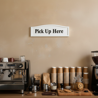 Coffee station with espresso machine, stacks of paper cups, and 'Pick Up Here' sign on beige wall.