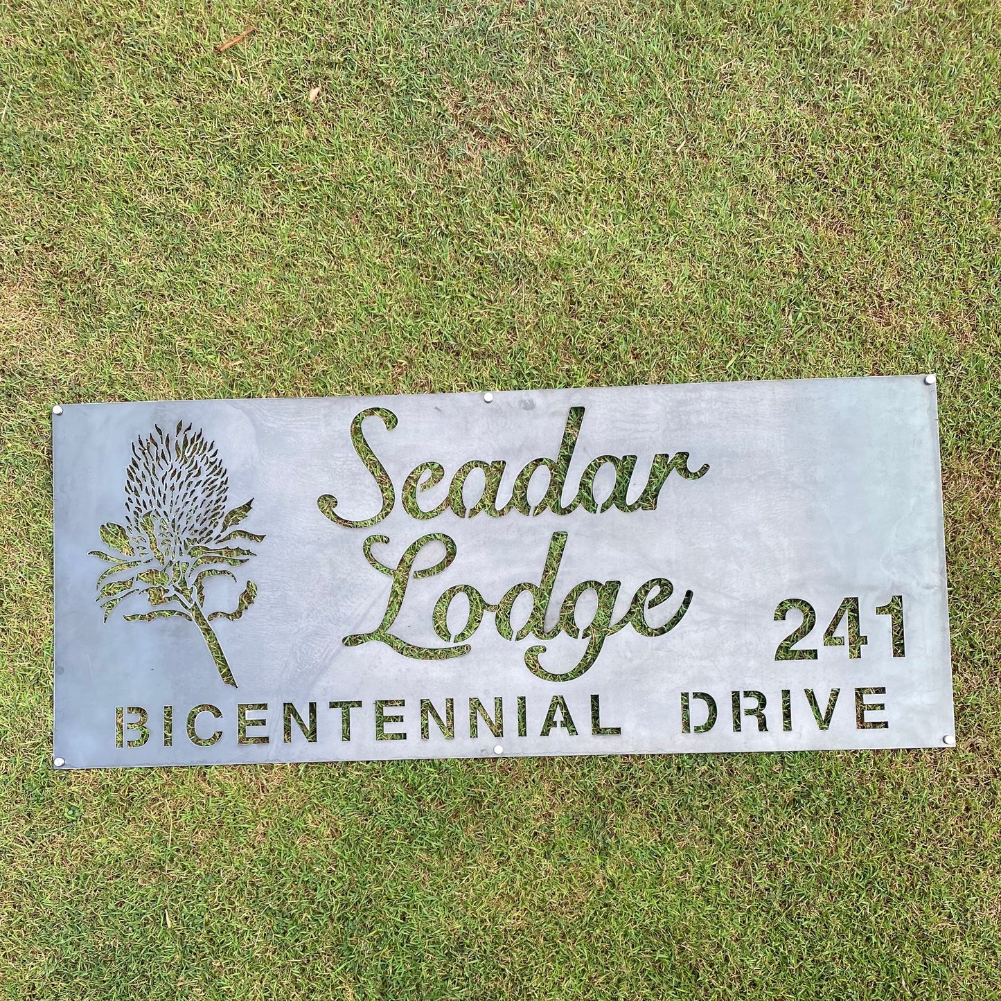 Metal sign for Seadar Lodge on Bicentennial Drive with a decorative plant design, placed on grass.