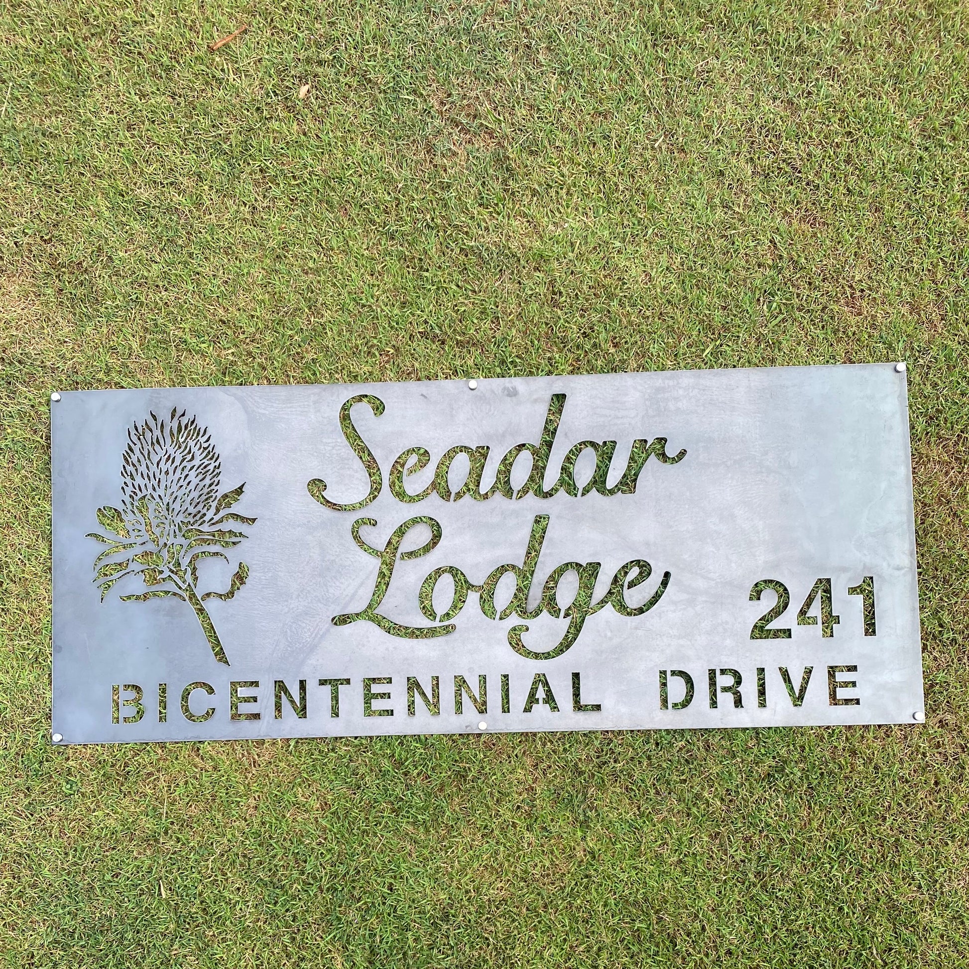 Metal sign for Seadar Lodge on Bicentennial Drive with a decorative plant design, placed on grass.